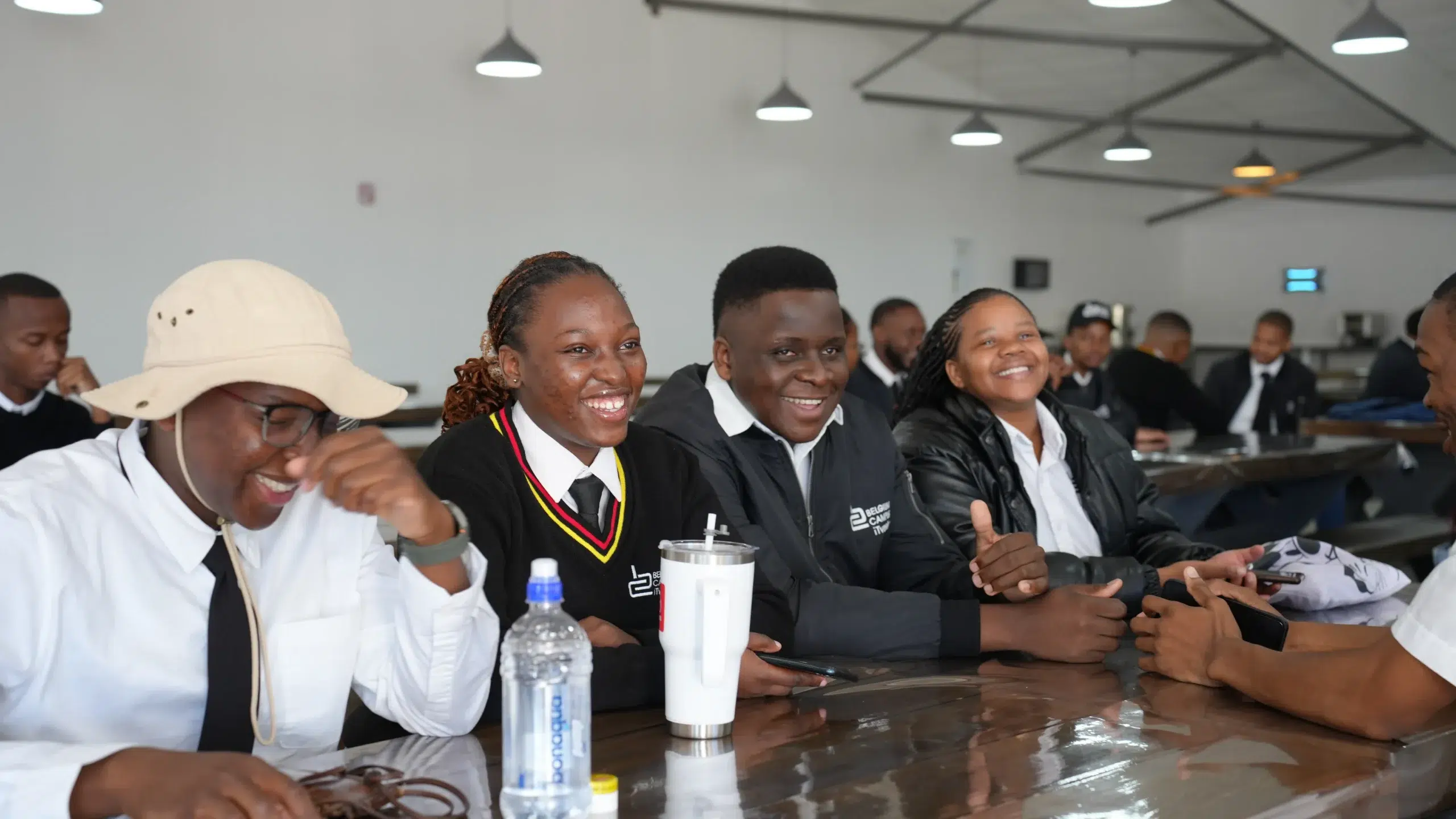 Group of students and young adults seated at a long table in a classroom, smiling and chatting during a meeting or class.