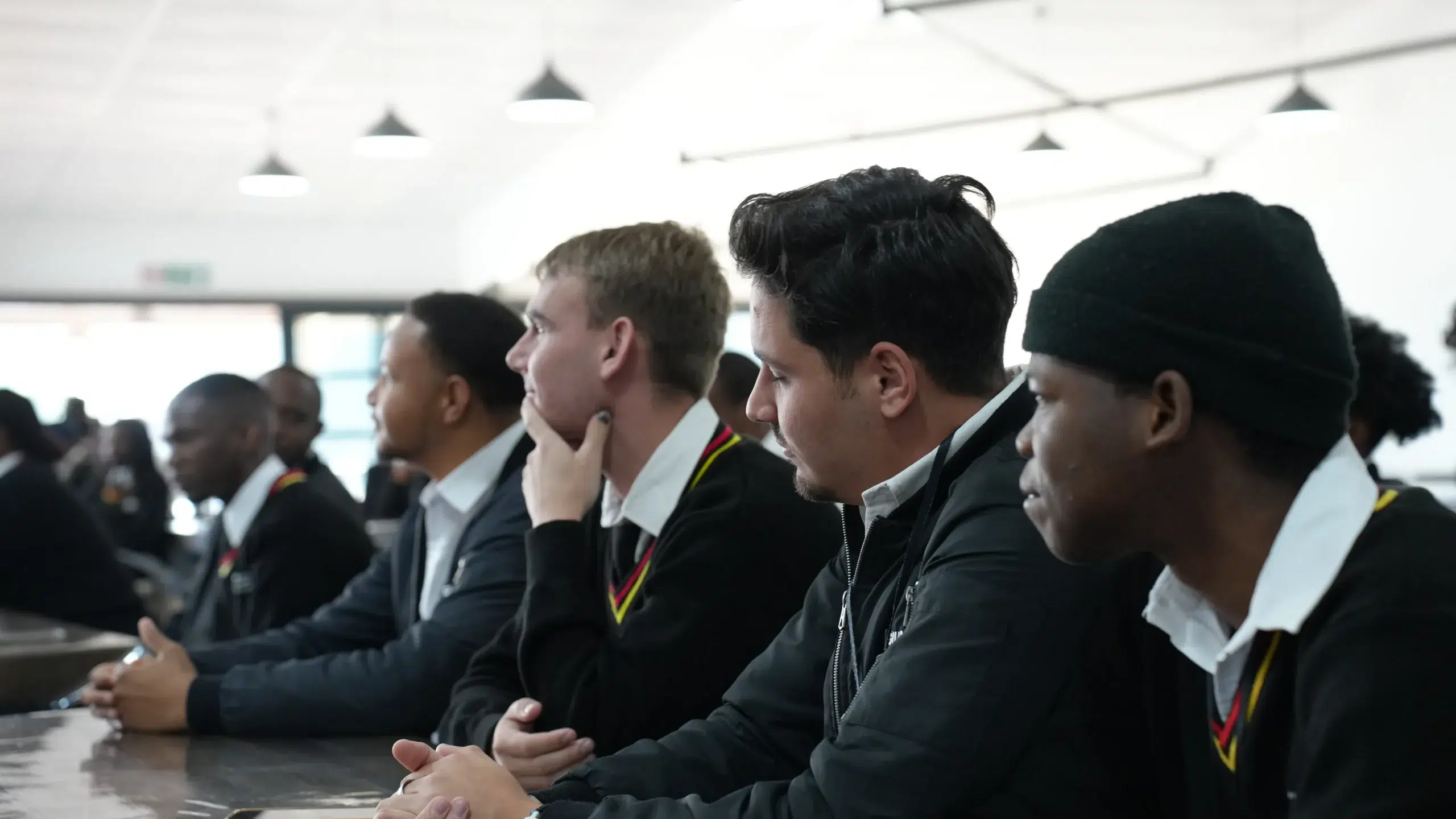 Group of students in matching uniforms seated at a long table in a bright classroom, listening attentively.