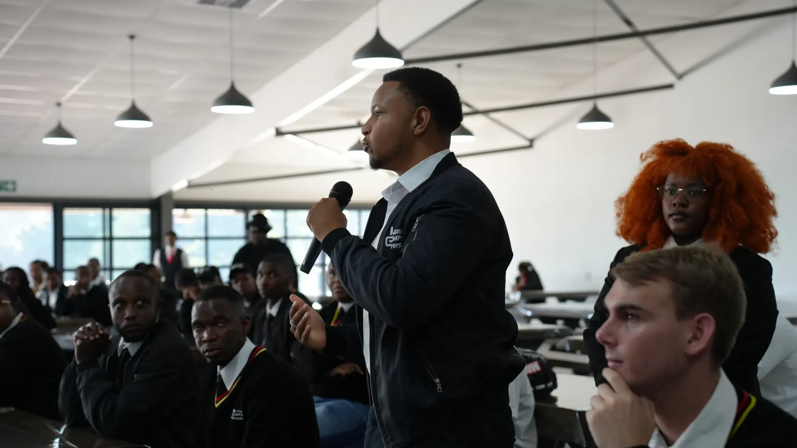 Young man in a dark jacket speaks into a handheld microphone to a seated student audience in a bright, modern hall.