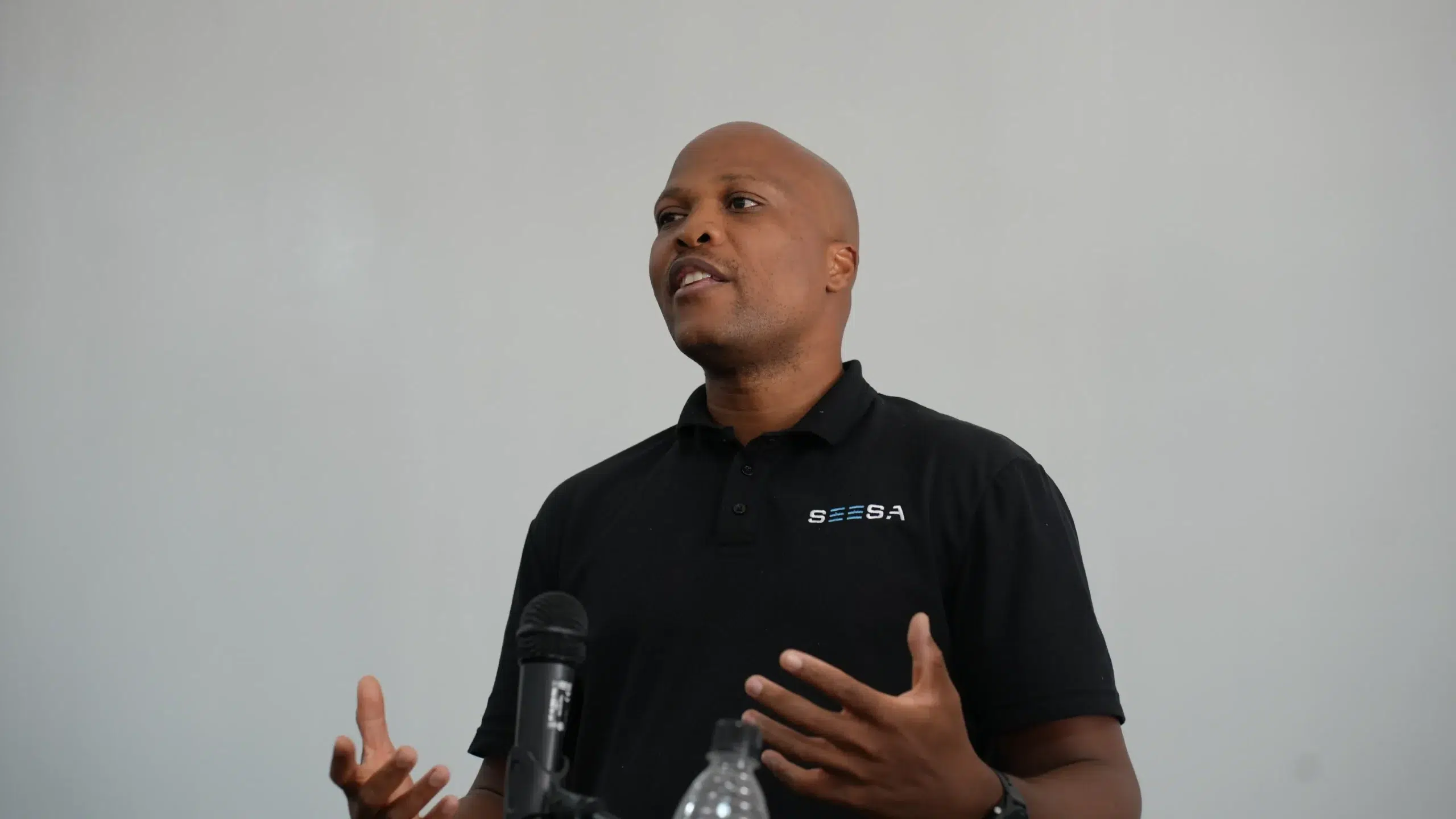 Mid-shot of a bald Black man in a black SEESA polo speaking into a microphone.