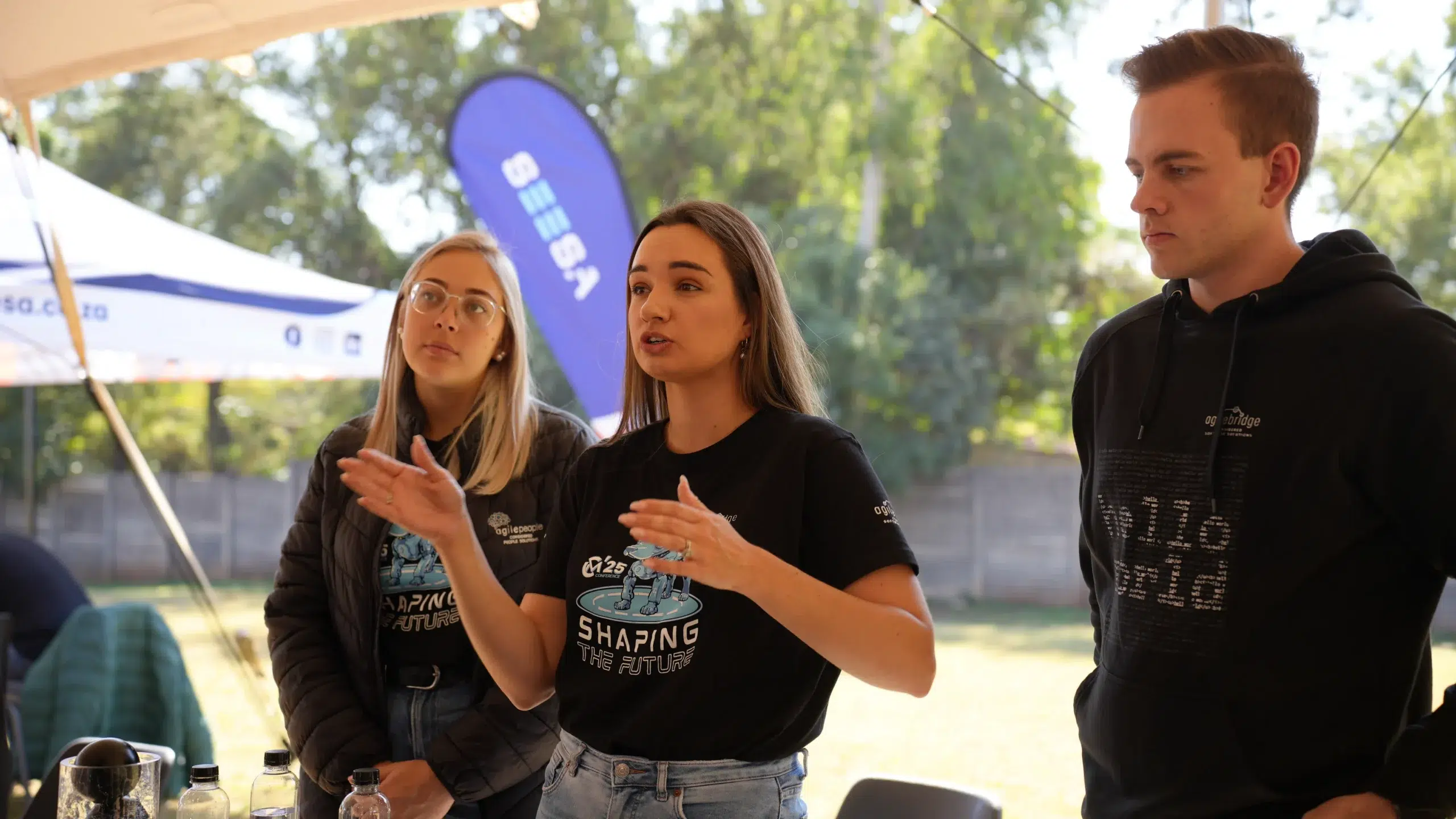 Three young adults standing outdoors, two women speaking while a man listens, at an event with tents and flags in the background.