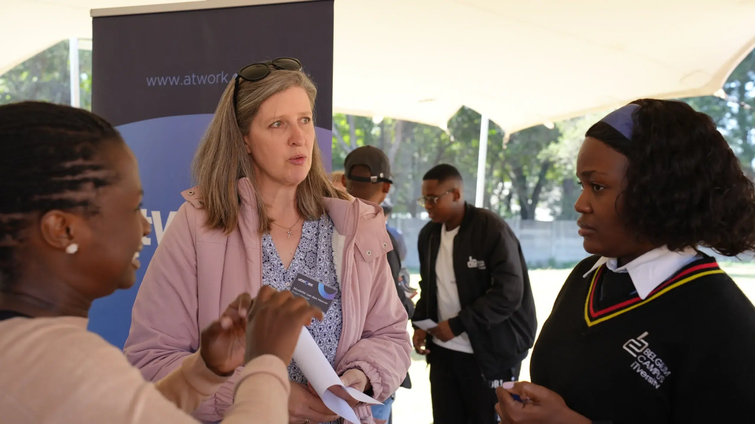 Group of people under a tent in a casual discussion; central woman in a pink jacket speaks while others listen.