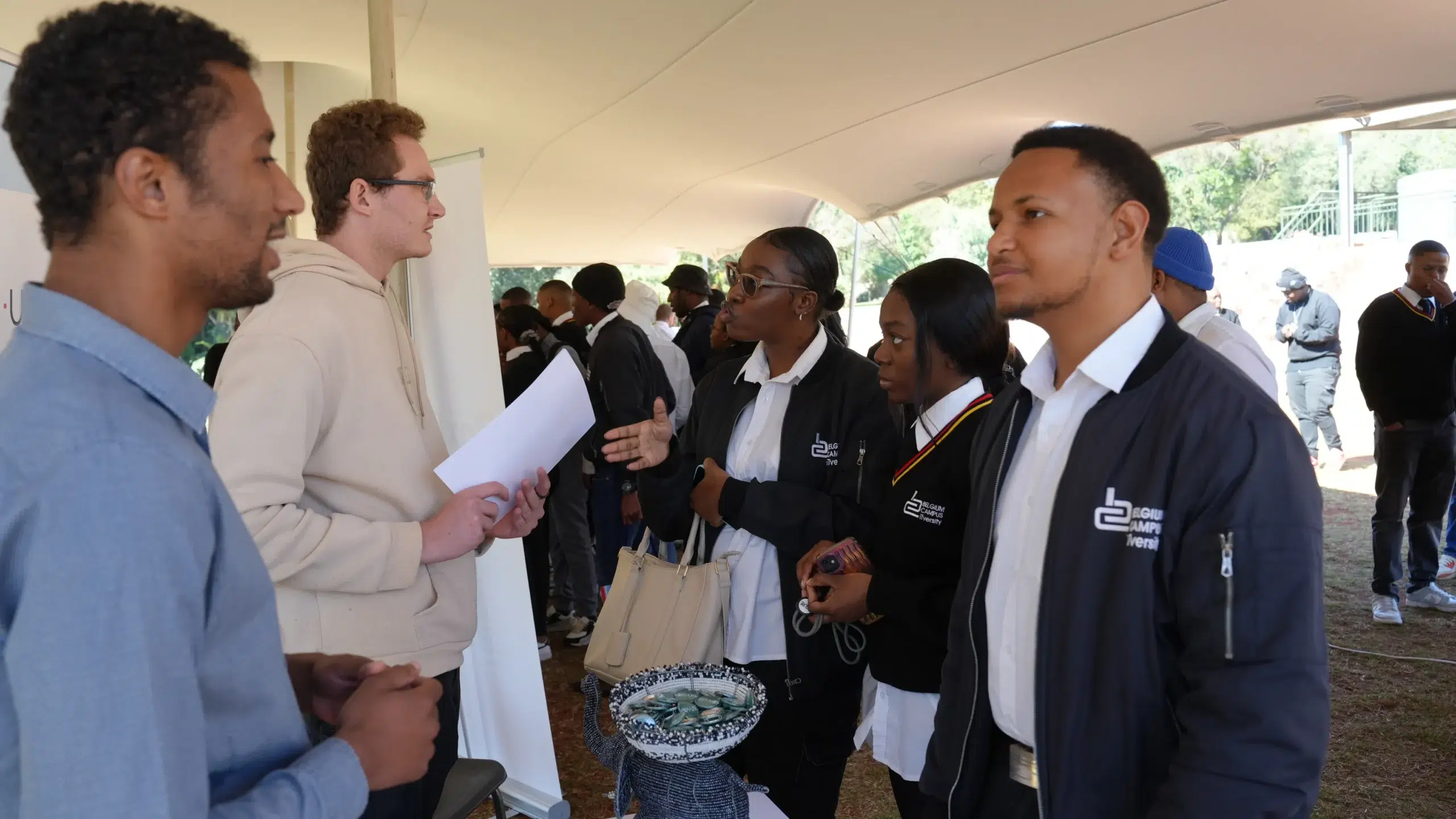 People handshake and chat under a white tent; students in uniforms share information with visitors, a basket of bracelets in foreground.