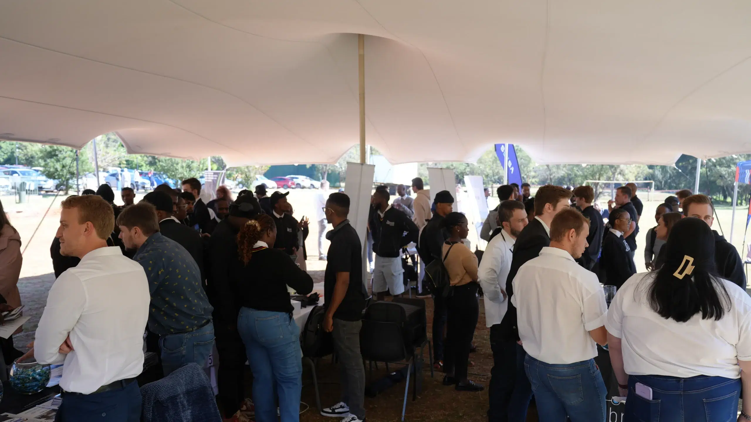 Crowded outdoor networking event under a large white tent with people chatting at booths under the canopy.
