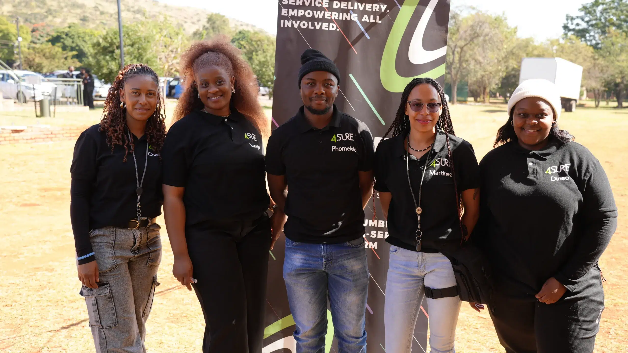Five people stand side by side outdoors, wearing black 4SURE polo shirts, posing in front of a tall event banner.