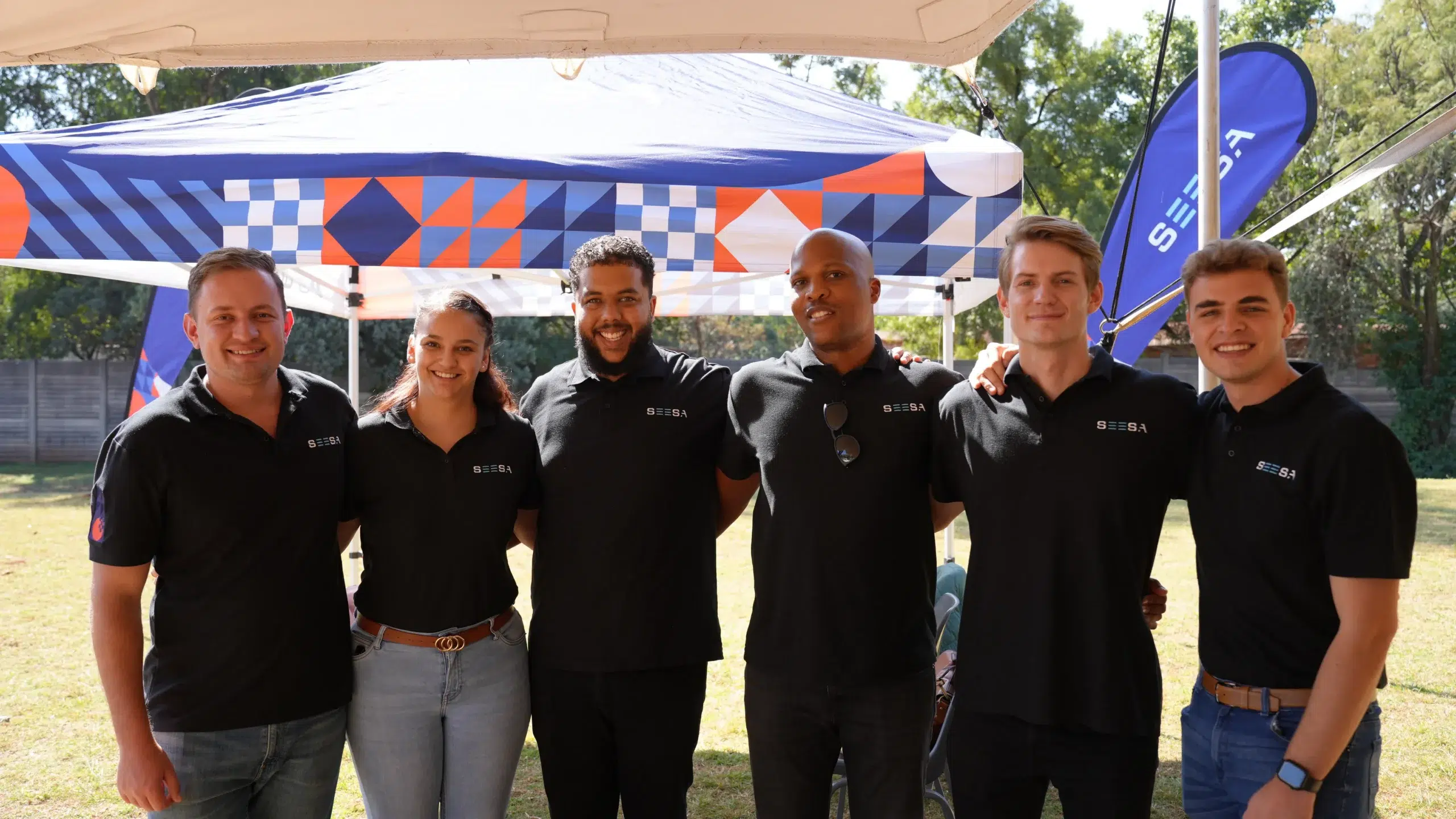 Six people in black polo shirts stand arm-in-arm under a patterned canopy at an outdoor event, smiling for the camera.