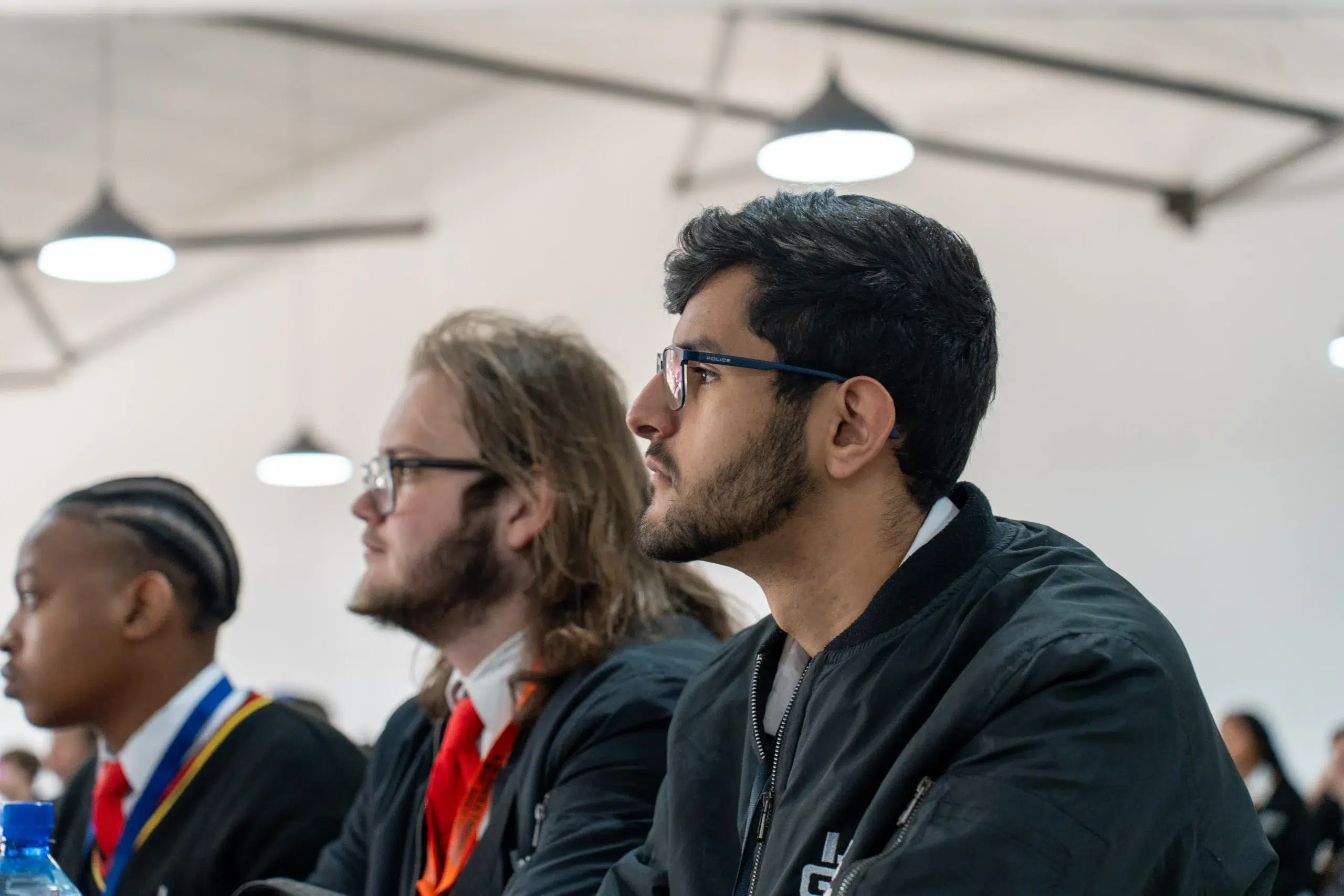 Side profile of a man with dark hair and blue glasses listening at a conference, wearing a black jacket.