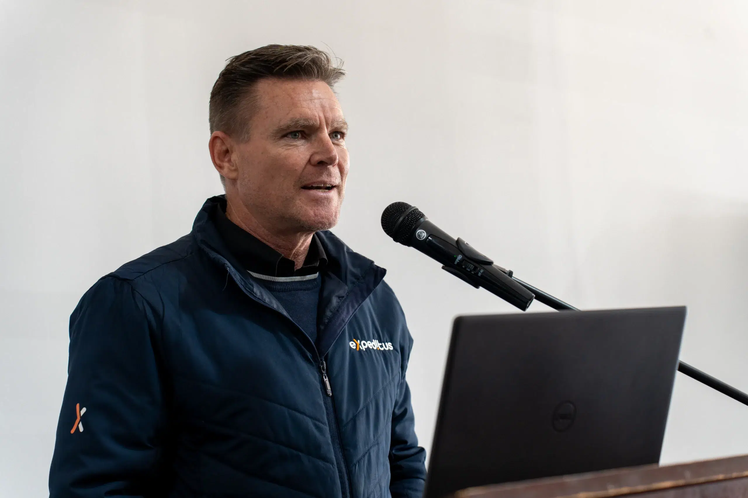 Male presenter speaking into a microphone at a podium, wearing a navy jacket with a small logo on the chest.