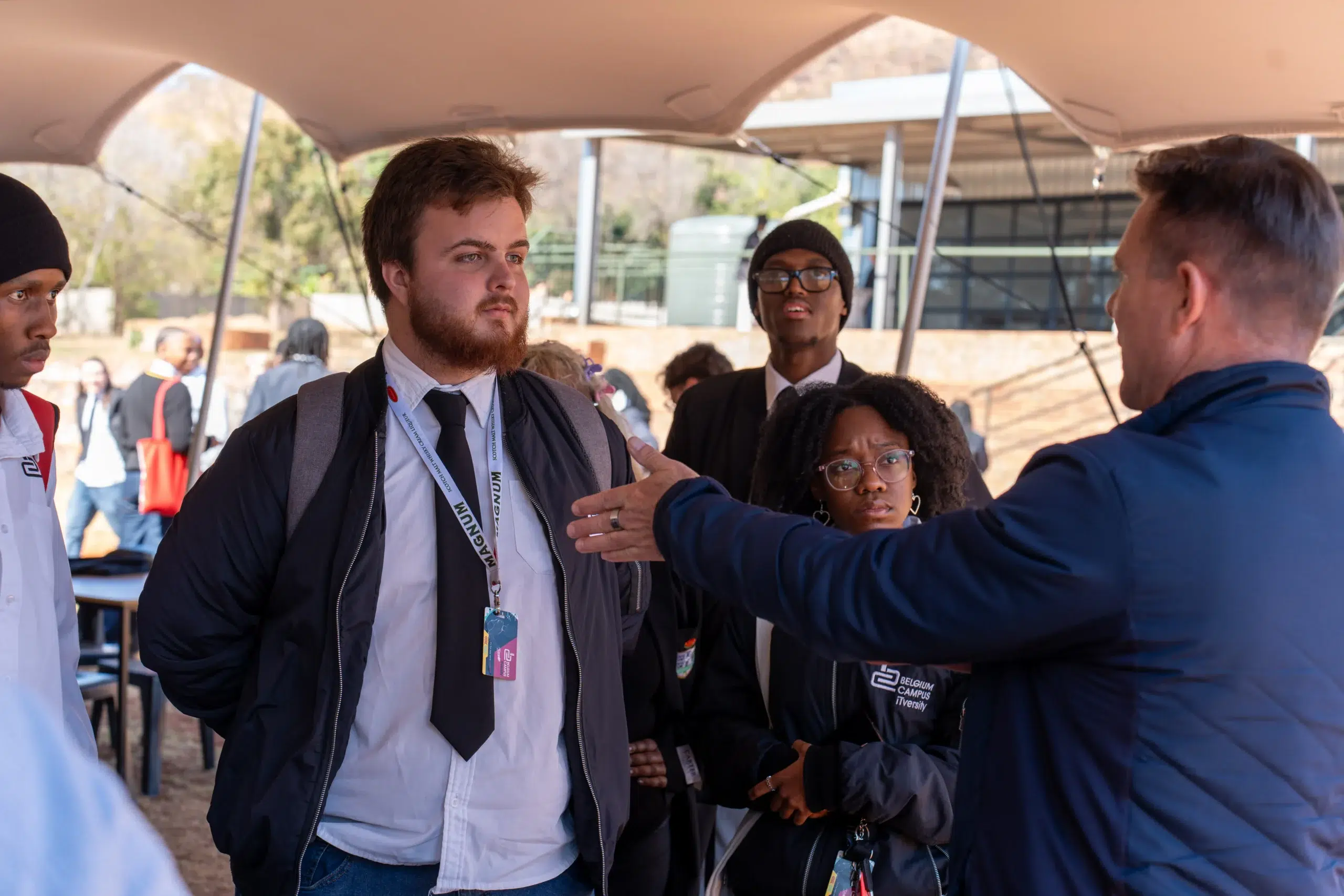 Group of students wearing lanyards and jackets talking under a canopy at an outdoor campus event.