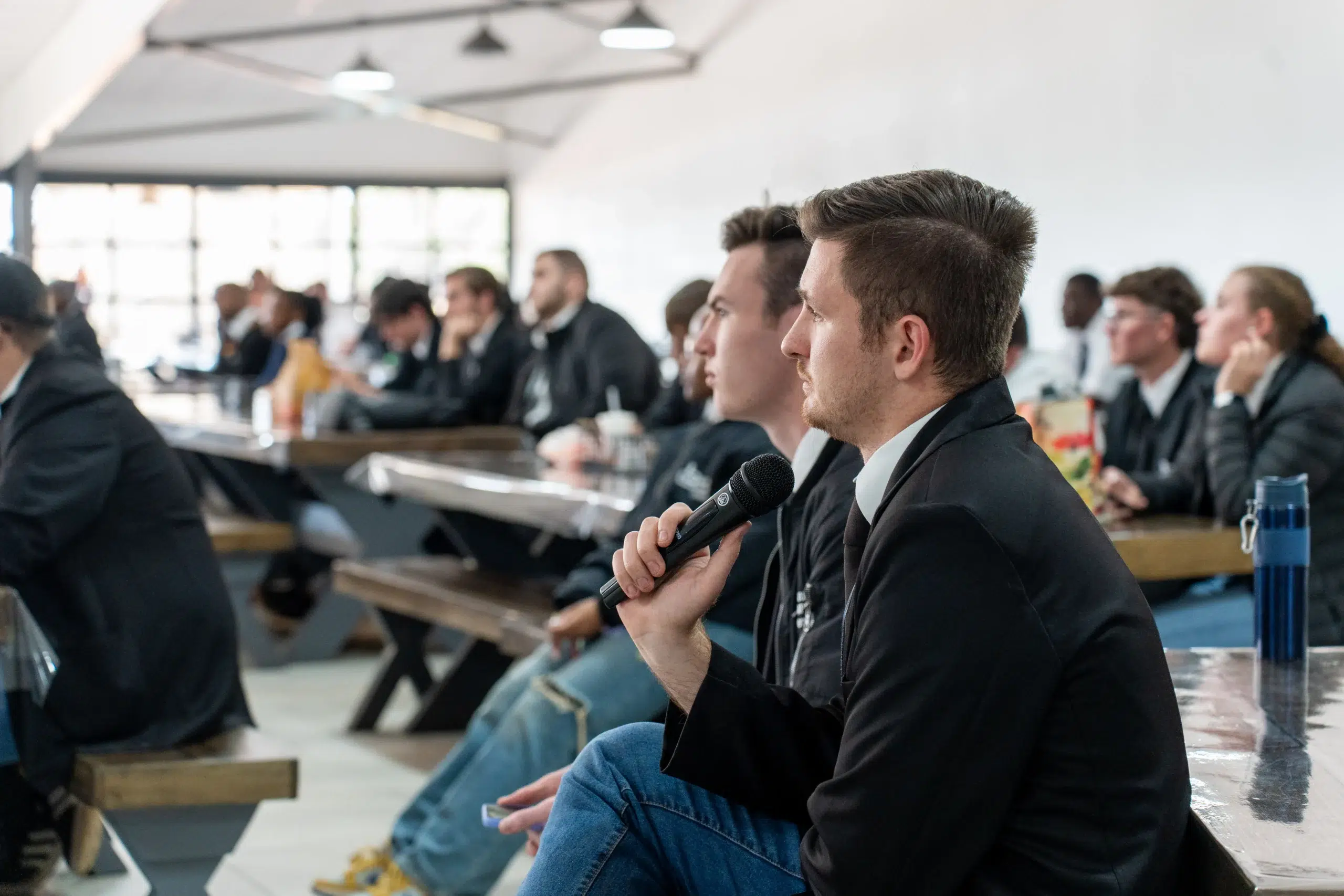 Man in the foreground wearing a blazer speaks into a handheld microphone during a meeting; others sit at desks in a bright classroom.issuing a question or comment in a conference.