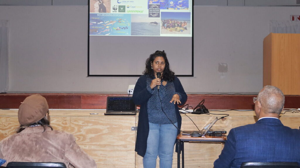 Woman stands on stage holding a microphone, giving a presentation with a projector screen behind her and an open laptop on a table nearby.