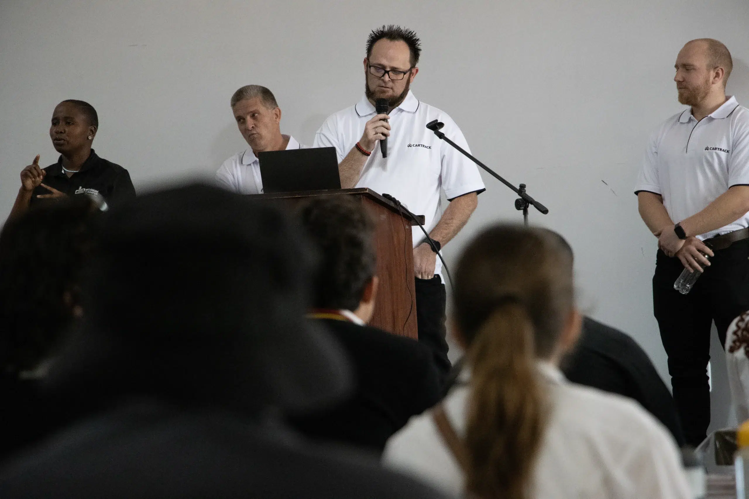 Group of four men on a stage; one man speaks into a handheld microphone at a wooden podium, the others stand nearby in white polo shirts.