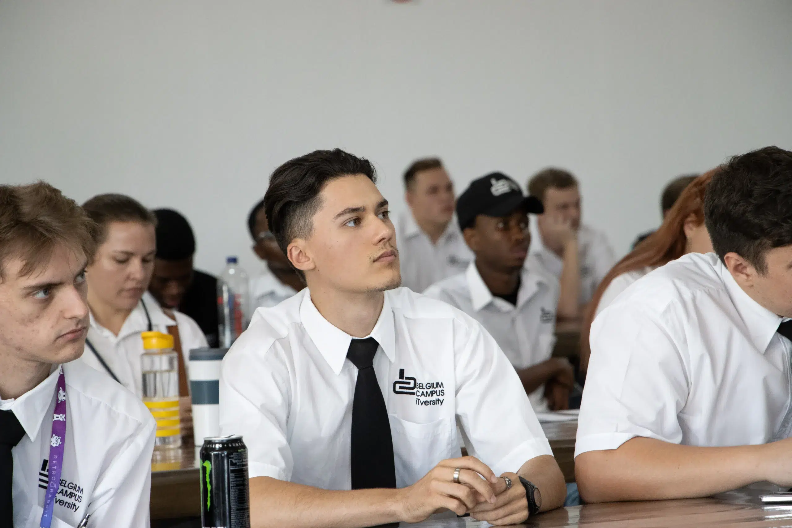 Group of students in white shirts seated at a classroom table, listening attentively; foreground student wears a black tie and a Belgium Campus University logo on his shirt.