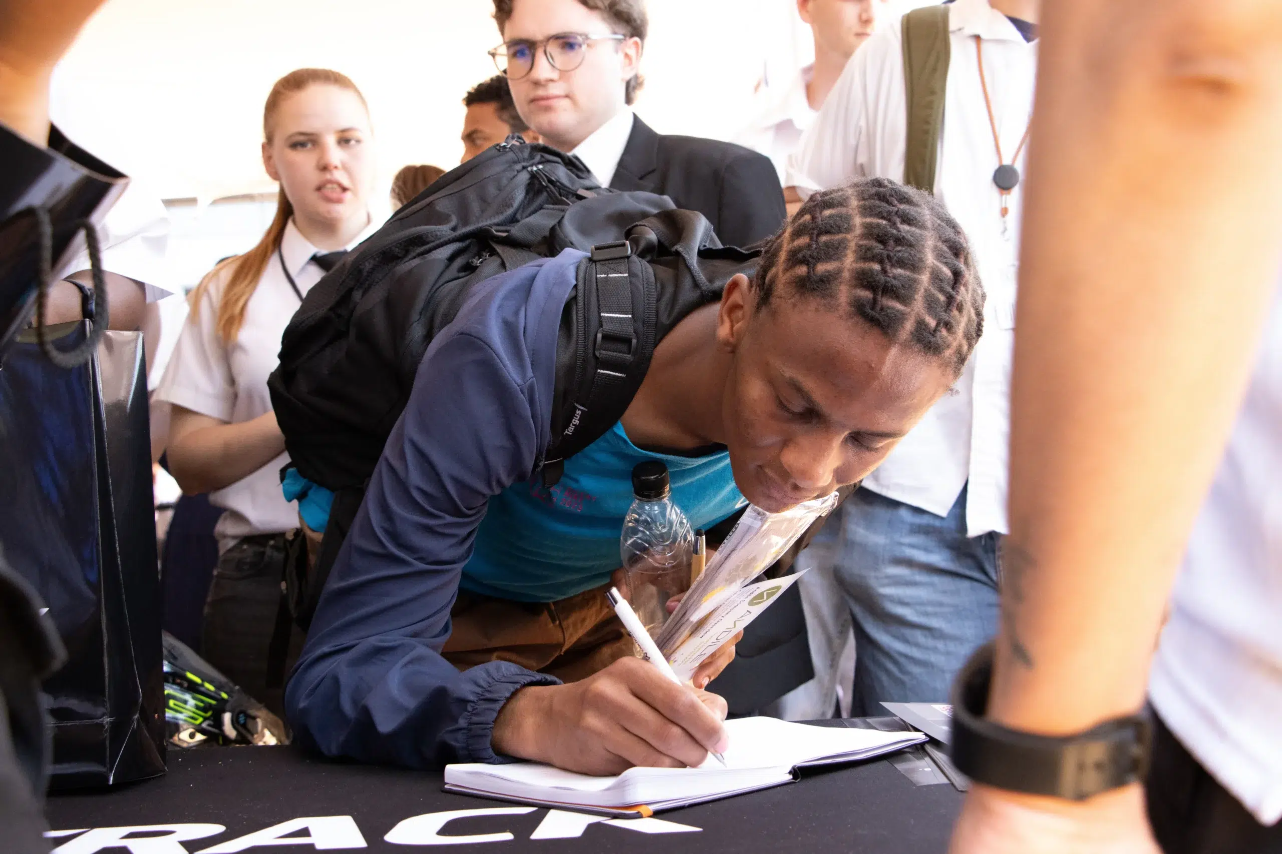 Student signs a notebook at a table while others stand nearby in a crowded event setting near a black tablecloth.