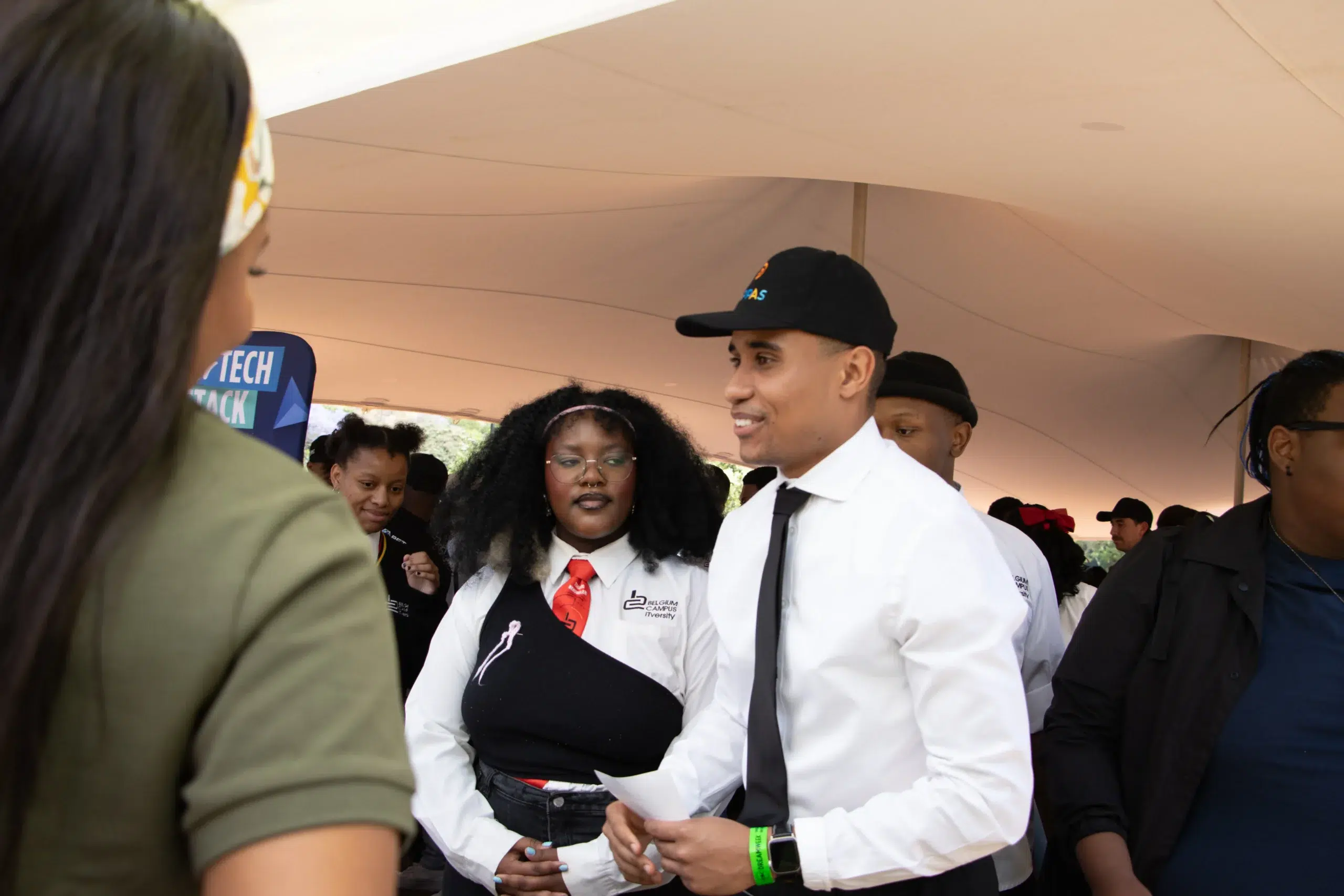 Group of young people at an outdoor event under a beige tent; man in white shirt and black tie smiles among attendees, holding papers.