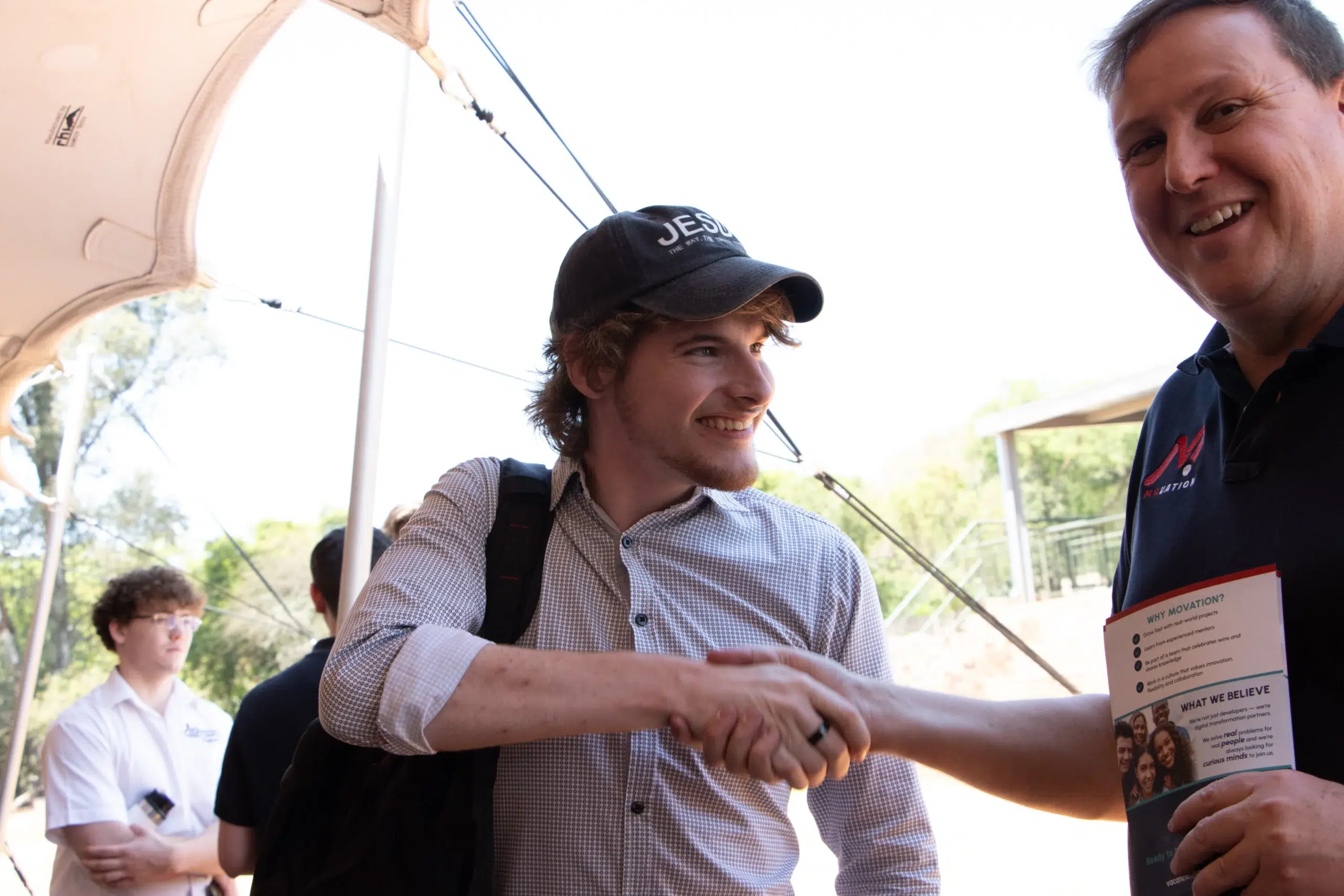 Two men shake hands and smile at an outdoor event, with a paper brochure visible in the foreground surface on the right margin.