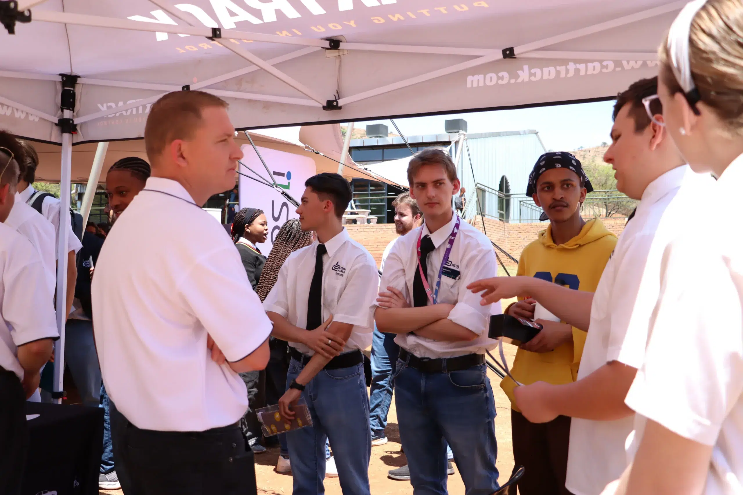 Group of students in white shirts and jeans under a canopy, chatting at an outdoor event; several wear lanyards and one person in a yellow hoodie stands nearby.