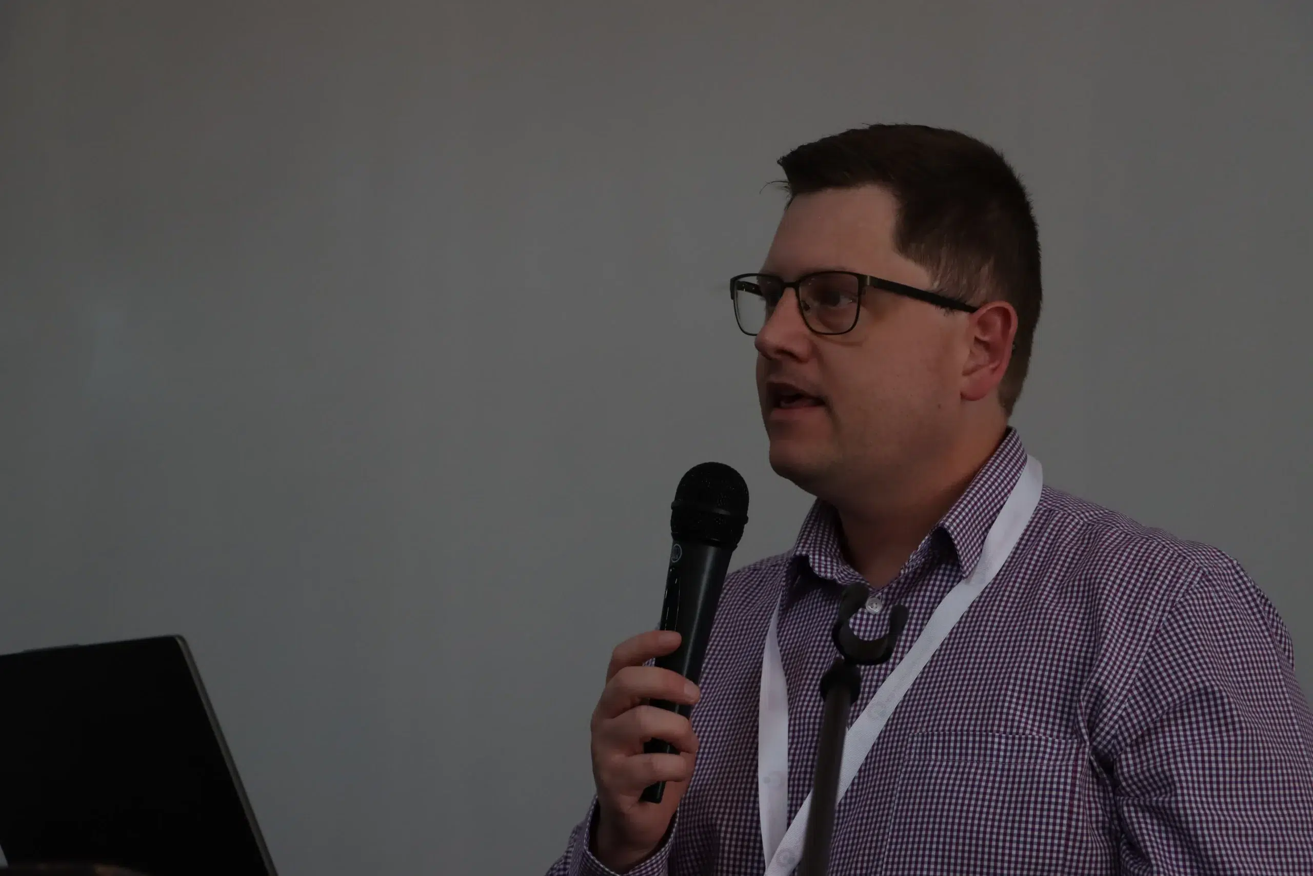 Man in glasses holding a microphone and speaking, wearing a checkered shirt and white lanyard at a conference or presentation.