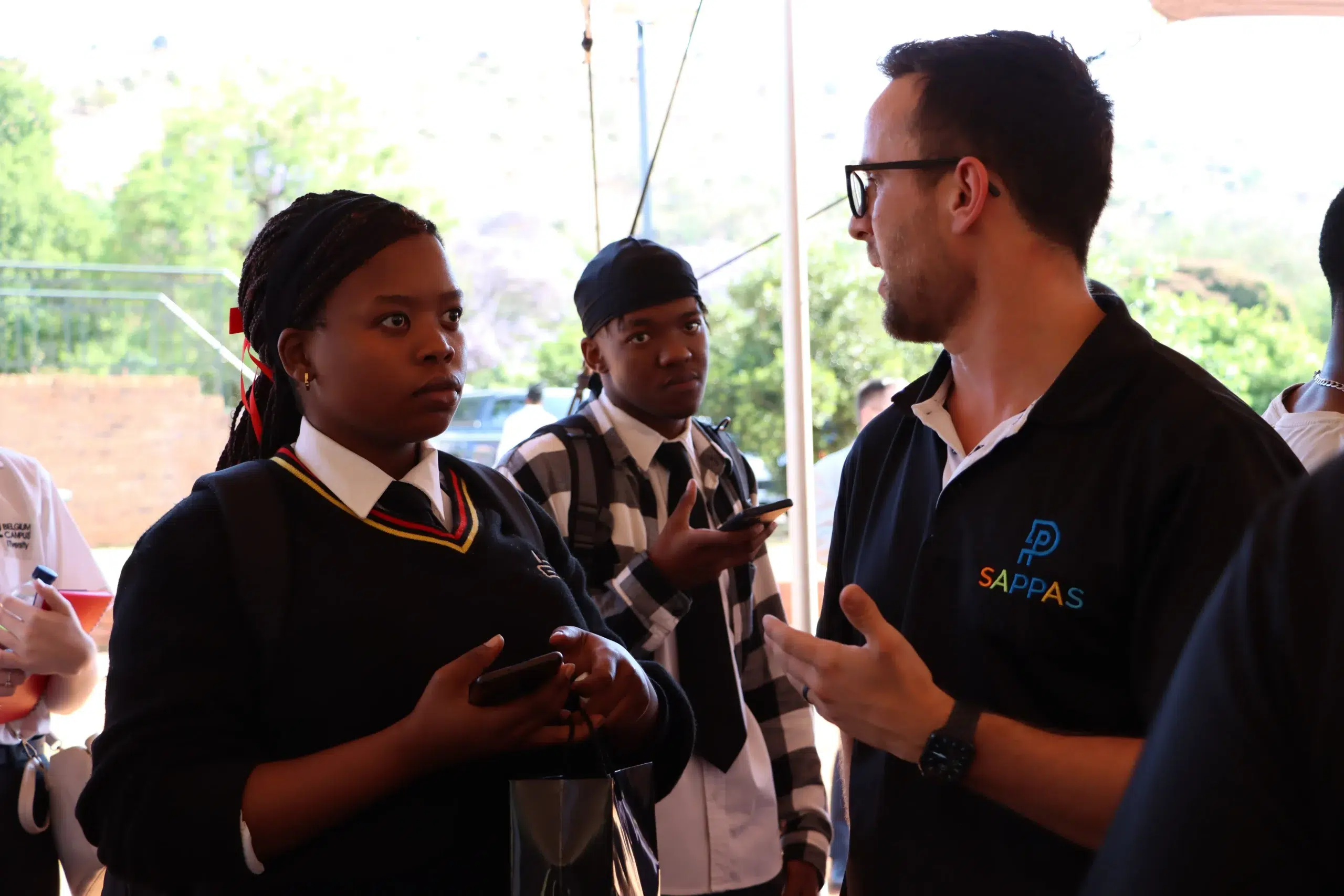 Group of students in uniform chat with a man wearing glasses at an outdoor campus event.