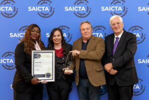 A group of four people stand in front of a blue SAICTA-branded backdrop. The person on the far left holds a framed certificate awarded to Jacqui Muller for winning the ICT Professional of the Year Award 2026. The second person from the left holds a glass trophy. All four individuals are smiling and posing for a celebratory photo.