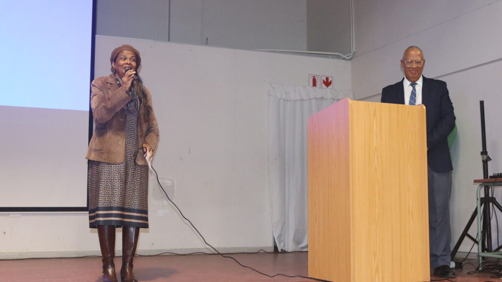 A woman speaks into a microphone on stage while another person stands at a podium beside her in a formal presentation setting, with a projection screen and exit sign behind them.