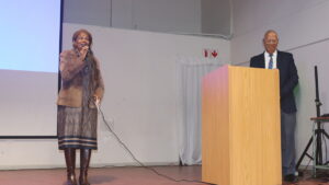 A woman speaks into a microphone on stage while another person stands at a podium beside her in a formal presentation setting, with a projection screen and exit sign behind them.