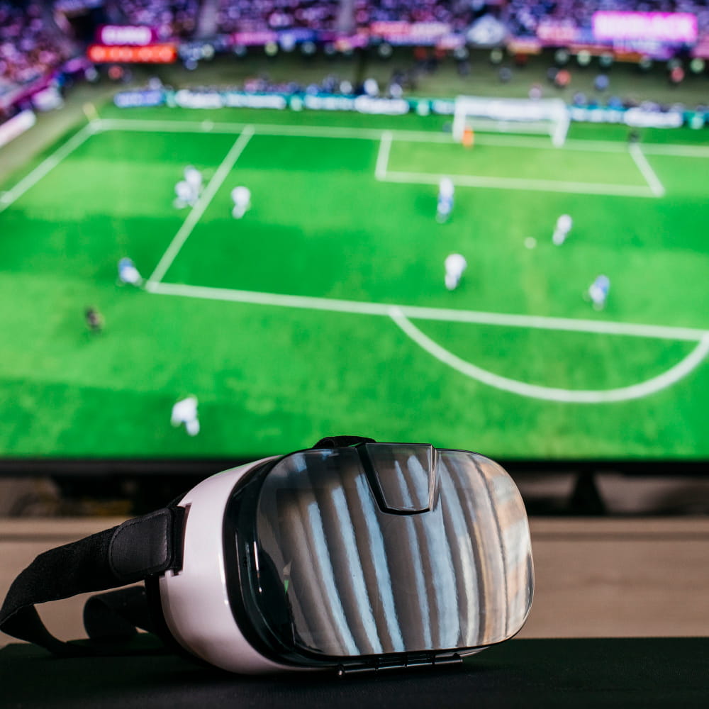 A virtual reality headset rests on a dark surface in the foreground, while a television in the background displays a live football match with players on a brightly lit green pitch.
