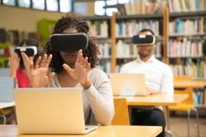 Three students sit at desks in a library, each wearing a virtual‑reality headset and interacting with digital content in the air. Laptops are open in front of them, and bookshelves line the background, illustrating a modern, technology‑enhanced learning environment.