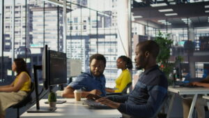 People working at desktop computers in a modern open-plan office with large floor-to-ceiling windows overlooking a cityscape. Multiple workstations are visible, with computer monitors, keyboards, and office desks arranged throughout a bright, professional workspace. Natural light fills the office, and indoor plants are visible in the background.