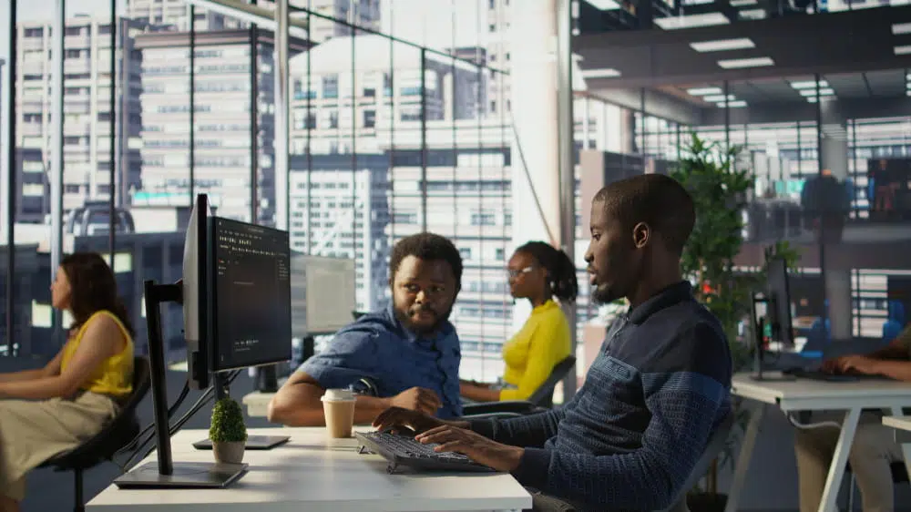 People working at desktop computers in a modern open-plan office with large floor-to-ceiling windows overlooking a cityscape. Multiple workstations are visible, with computer monitors, keyboards, and office desks arranged throughout a bright, professional workspace. Natural light fills the office, and indoor plants are visible in the background.
