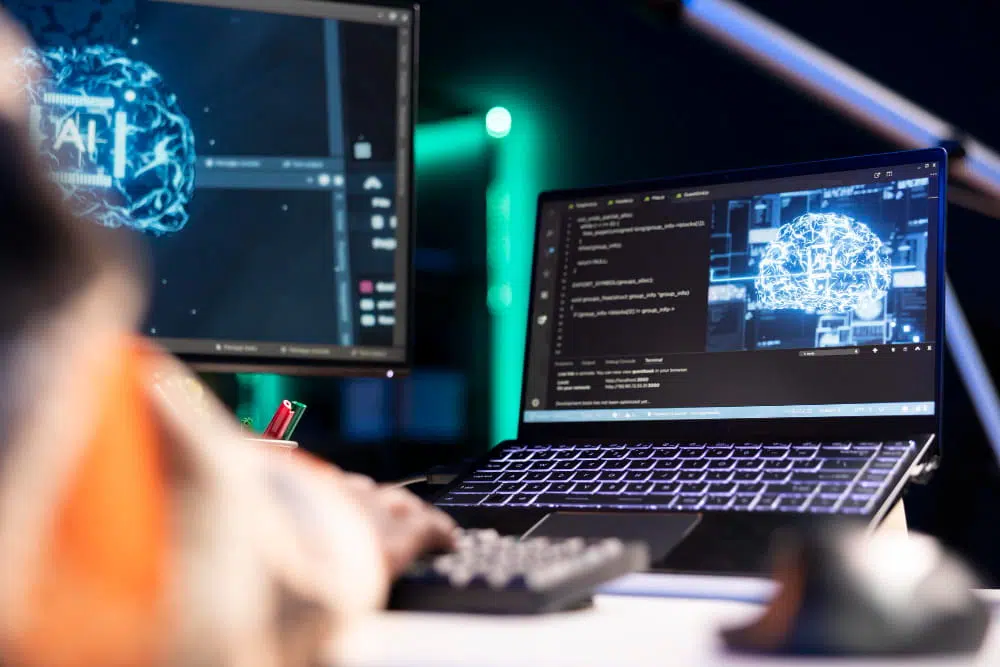 A person works at a desk with two computer screens displaying glowing digital brain graphics symbolising artificial intelligence. The left monitor shows a circuit‑patterned brain with the letters “AI” inside it, while the laptop screen shows a similar brain alongside lines of code. The person’s hands rest on the keyboard, and a mouse and pen holder sit nearby. The scene is lit with blue and green tones, creating a high‑tech atmosphere.