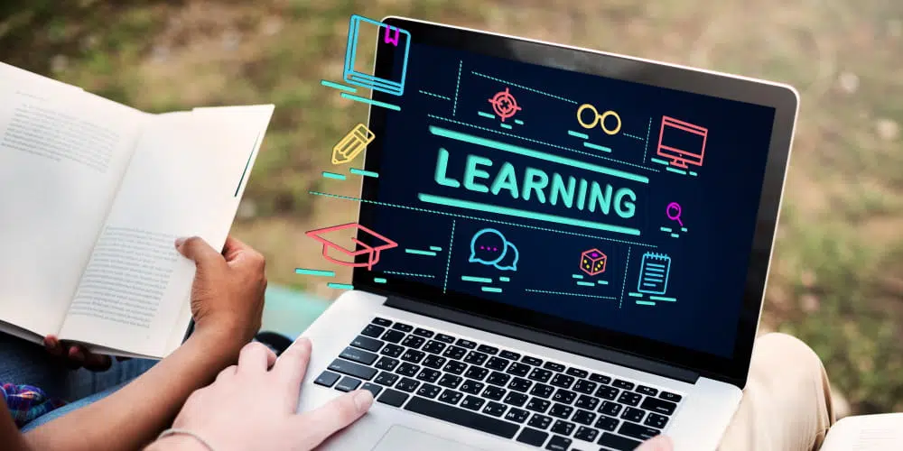 A person sits outdoors holding an open book next to a laptop. The laptop screen displays the word “LEARNING” in bright letters surrounded by colourful education‑themed icons, including a graduation cap, pencil, speech bubble, magnifying glass, computer monitor, and notepad. The scene highlights a blend of digital and traditional study methods.