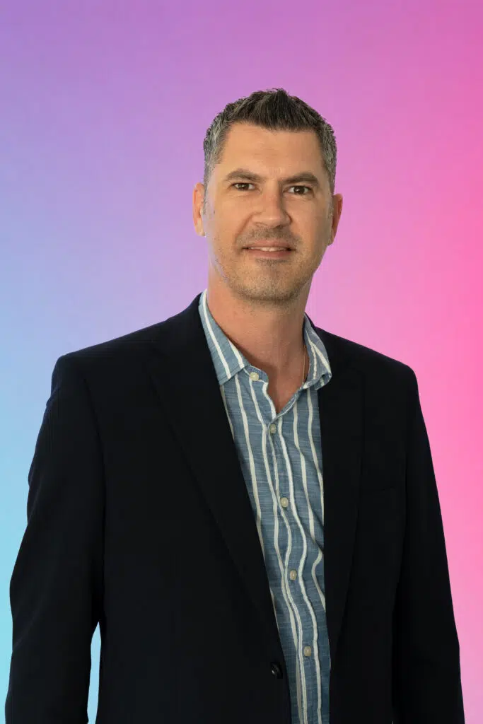 Portrait of a man in a dark blazer and blue striped shirt, posed against a pink-to-blue gradient background.
