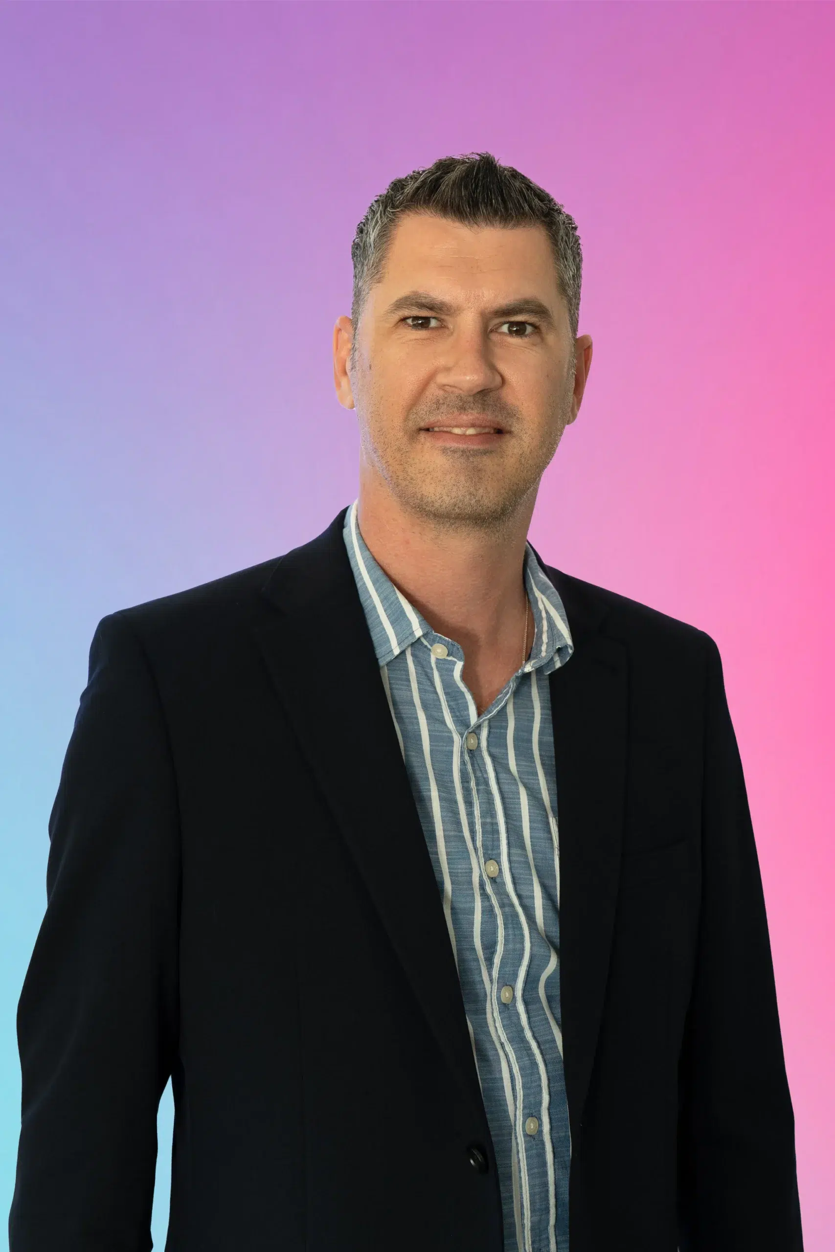 Portrait of a smiling man in a navy blazer and striped shirt against a pink‑to‑blue gradient background.