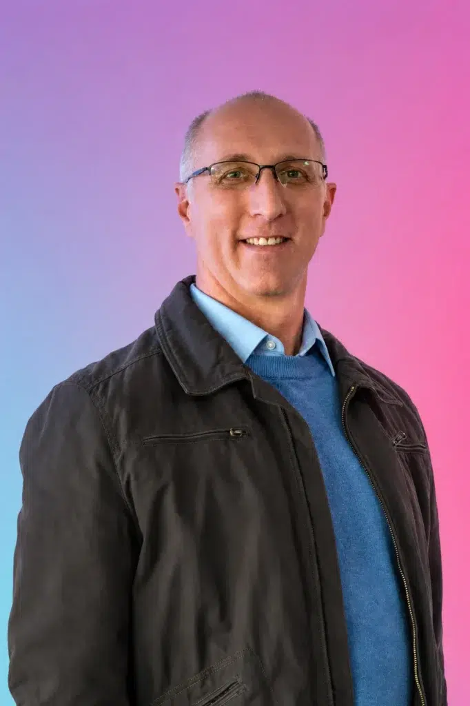 Portrait of a smiling middle-aged man wearing glasses, a blue sweater, and a light blue collared shirt, against a pink-to-blue gradient background.