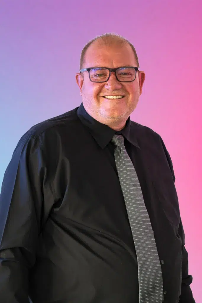 Smiling man in a black shirt with a gray dotted tie, against a pink-to-blue gradient background.