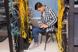 A person sits on a chair between two tall server racks filled with neatly organised yellow and blue network cables. They are wearing a checkered shirt, jeans, and grey shoes, and are working on a laptop. The scene takes place in a server room or data centre, highlighting hands‑on network maintenance and the complexity of modern digital infrastructure. Network engineering courses