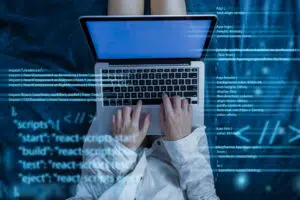 A person sits on a blue surface using a laptop, typing with both hands on the keyboard. Overlaid across the image are translucent lines of JavaScript and CSS code, suggesting programming or web development activity.