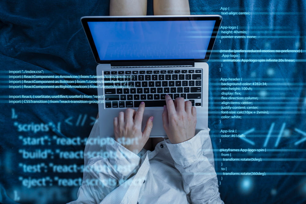 A person sits on a blue surface using a laptop, typing with both hands on the keyboard. Overlaid across the image are translucent lines of JavaScript and CSS code, suggesting programming or web development activity.