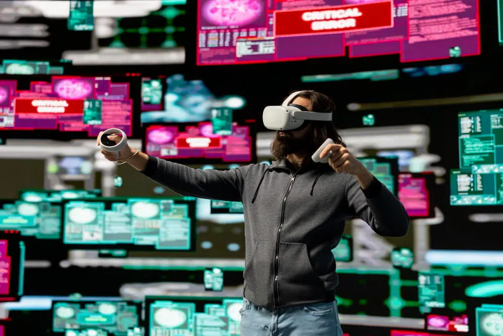 A person wearing a virtual reality headset stands in front of multiple floating digital screens filled with colourful interface elements. They hold two VR controllers while several panels display warnings such as “CRITICAL ERROR,” creating the impression of navigating a complex virtual data environment.