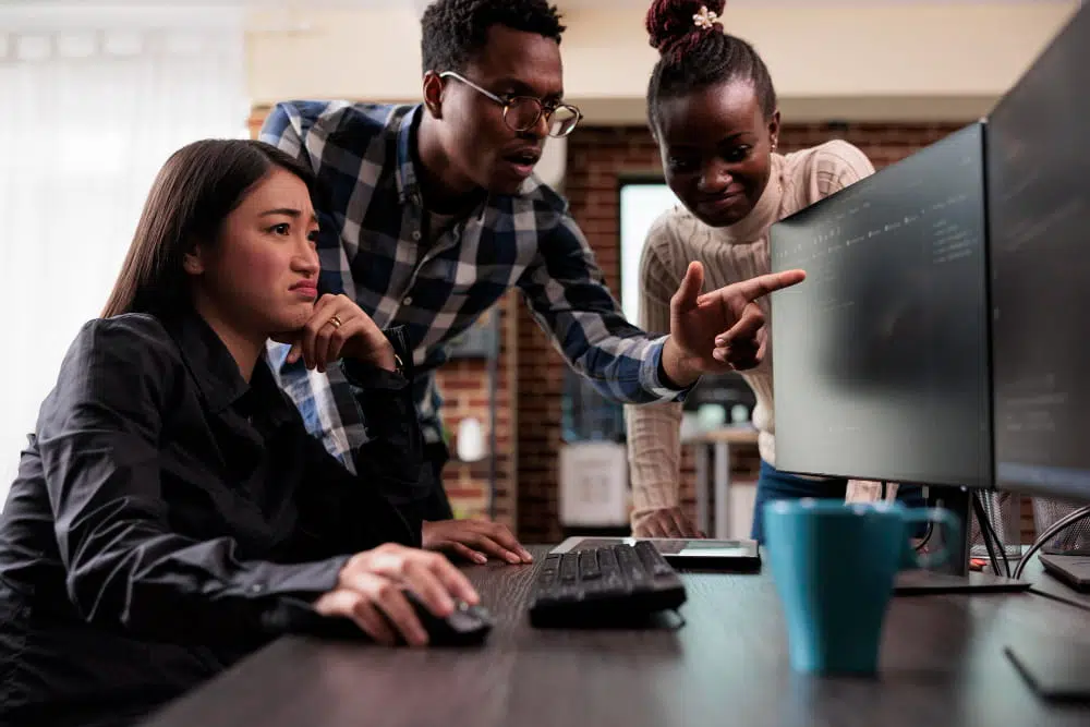 Three people are gathered around a desk in an office workspace, looking at a large computer monitor. One person is seated using a mouse and keyboard, while two others stand nearby, with one pointing at information displayed on the screen. The desk contains a keyboard, mouse, and a blue cup. The background includes brick walls and office furnishings, and the scene appears to show a collaborative discussion around computer-based work.
