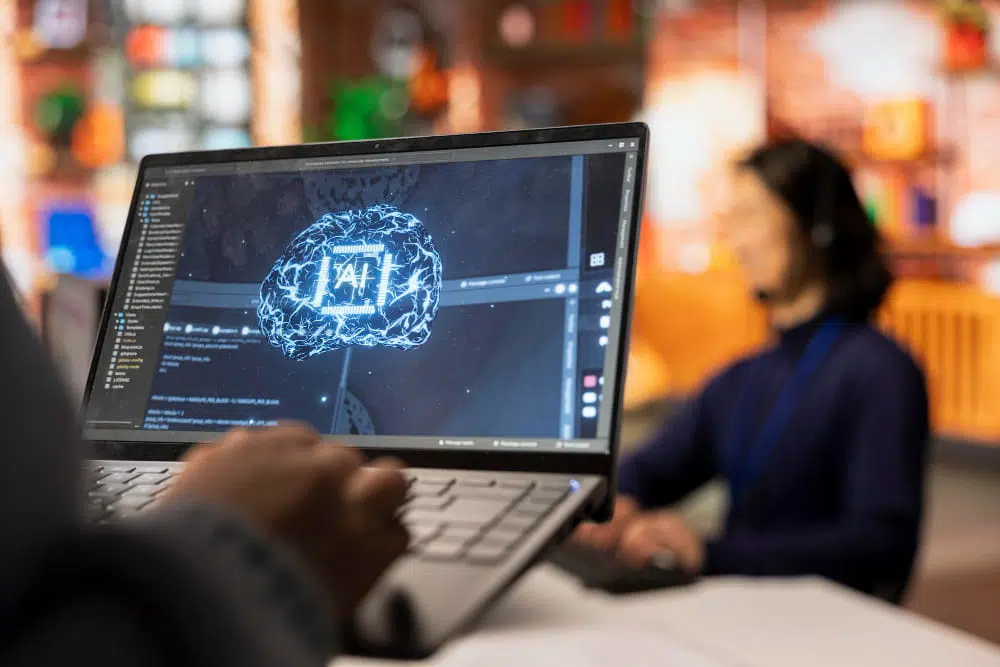 A close-up of a laptop screen displaying a digital illustration of a brain made of circuitry with the letters “AI” in the centre. Lines of programming code and a development interface appear in the background of the screen. Behind the laptop, a person wearing a headset sits in a warm, modern workspace with shelves and soft lighting. Generative AI