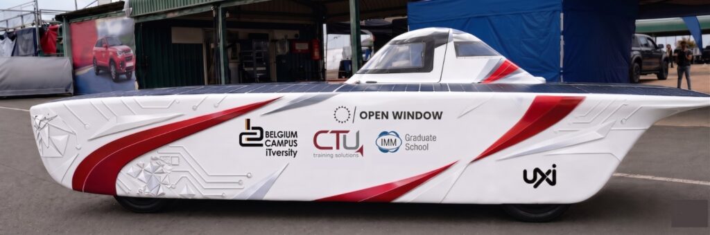 A white solar-powered vehicle with solar panels mounted across its top is parked in front of a paddock at a racing circuit. The vehicle displays logos for Open Window, UXI, Belgium Campus iTversity, CTU Training Solutions, and IMM Graduate School, with the number 60 visible on the side. A blue tent labelled "Solar Challenge Team" stands behind the vehicle, alongside covered pit garages.