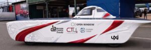 A white solar-powered vehicle with solar panels mounted across its top is parked in front of a paddock at a racing circuit. The vehicle displays logos for Open Window, UXI, Belgium Campus iTversity, CTU Training Solutions, and IMM Graduate School, with the number 60 visible on the side. A blue tent labelled "Solar Challenge Team" stands behind the vehicle, alongside covered pit garages.