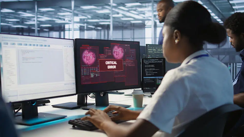 Open-plan office with several monitors; the central screen shows a red 'CRITICAL ERROR' alert as developers work nearby.