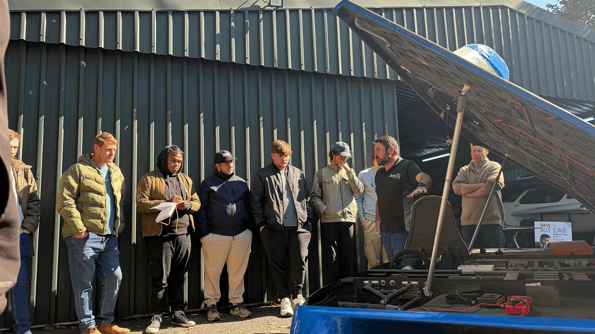 A group of students stand outdoors beside a blue solar-powered vehicle while a presenter explains its internal components. The vehicle's upper panel is raised, revealing wiring and technical equipment, with a globe mounted above the structure. The demonstration takes place outside a green corrugated-metal building in bright daylight.
