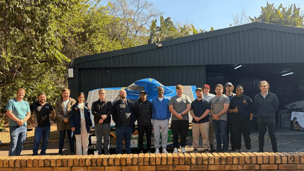 A group poses for a photo in front of a solar-powered vehicle outside a large workshop building. The vehicle is covered with solar panels, and trees surround the site on a sunny day.
