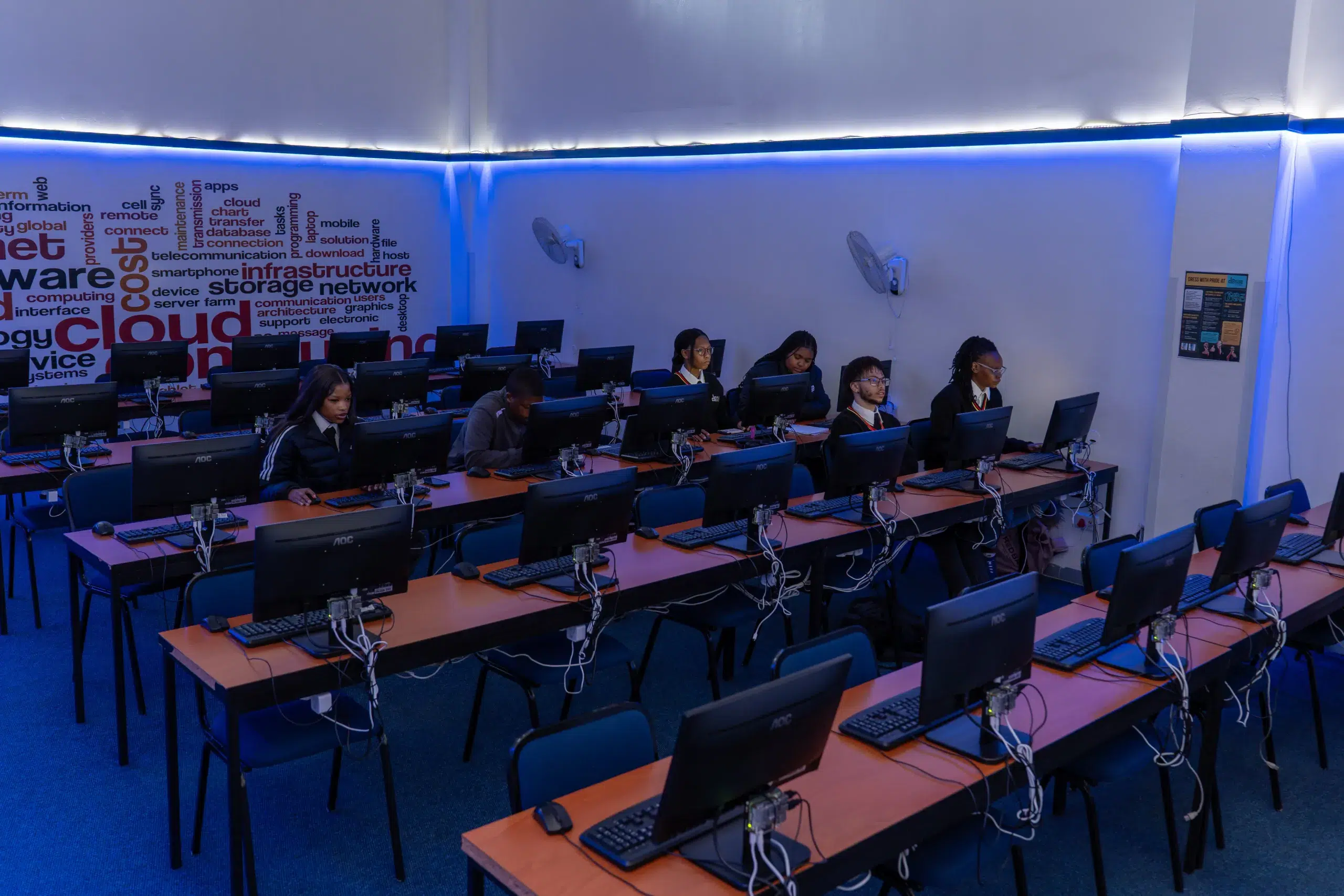 Computer lab with students at desks using desktop PCs, blue ambient lighting and a wall graphic of tech terms in the background.