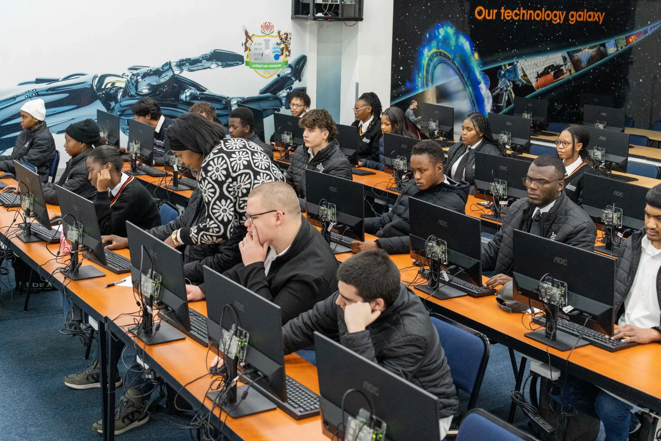 Group of students working on desktop computers in a computer lab with orange tables and a space-themed mural in the background