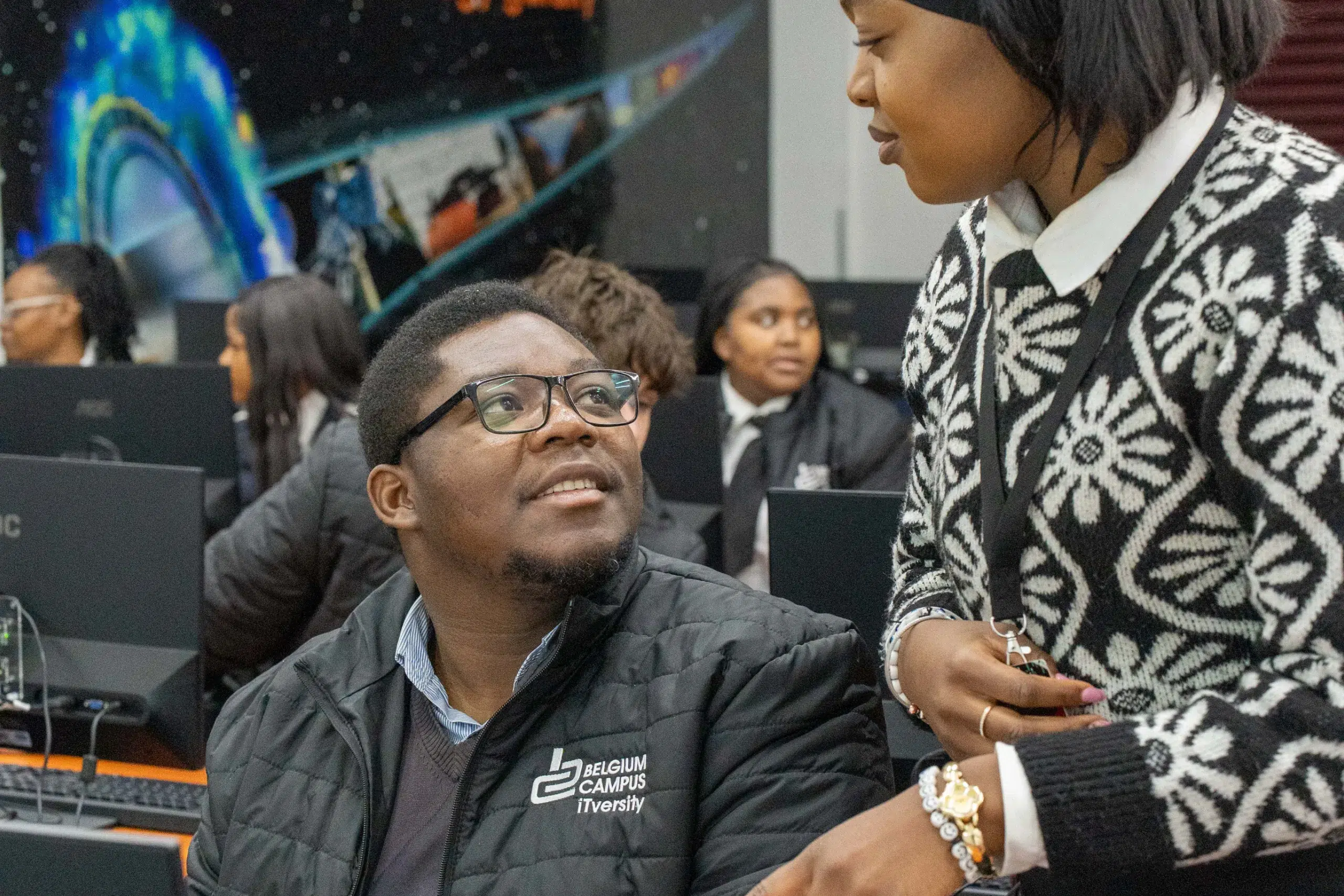 Male student with glasses sits at a computer in a campus lab, looking up at a standing woman in a patterned sweater.